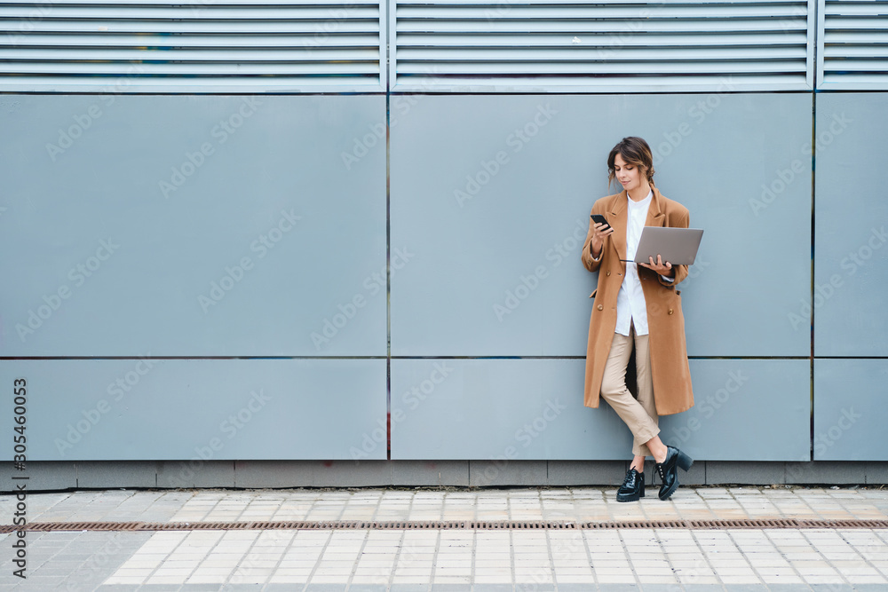 Young businesswoman in coat with laptop using cellphone outdoor