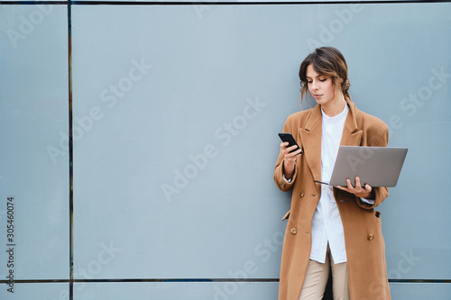 Young beautiful businesswoman in coat with laptop confidently using cellphone outdoor
