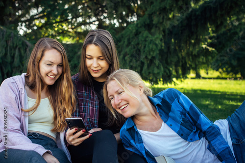 Three young pretty women on the lawn in park look at smartphone and laugh. Female students relax in the park sitting on green grass. Blonde, brunette and brown-haired outdoor, close-up.