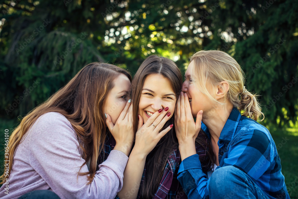 Three young attractive woman sharing secrets sitting on green grass in the park. Cheerful girlfriends gossip and whisper outdoor.