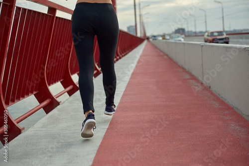 Girl athlete is jogging in the morning outside, in the city, on modern bridge crossing river. Active healthy lifestyle. Back view.