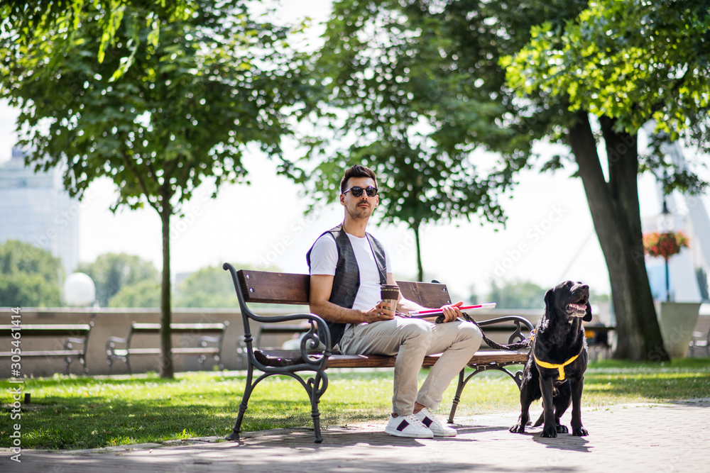 Young blind man with white cane and guide dog sitting in park in city. Young blind man with white cane and guide dog sitting in park in city.
