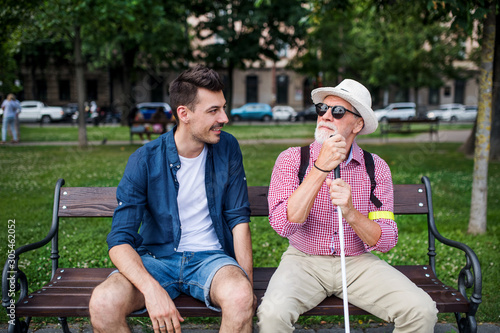 Young man and blind senior with white cane sitting on bench in park in city.