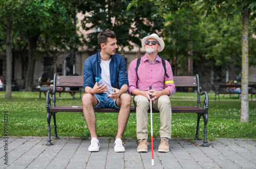 Young man and blind senior with white cane sitting on bench in park in city.