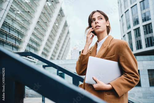 Young pensive casual businesswoman in coat with laptop talking on cellphone thoughtfully looking away outdoor