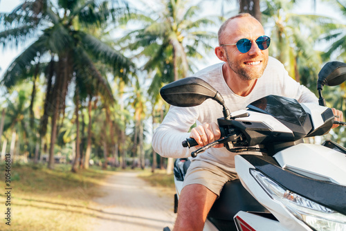 Happy smiling male tourist in sunglasses riding motorbike scooter during his tropical vacation under palm trees.