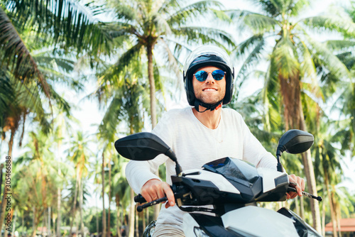 Happy smiling male tourist in helmet and sunglasses riding motorbike scooter during his tropical vacation under palm trees.