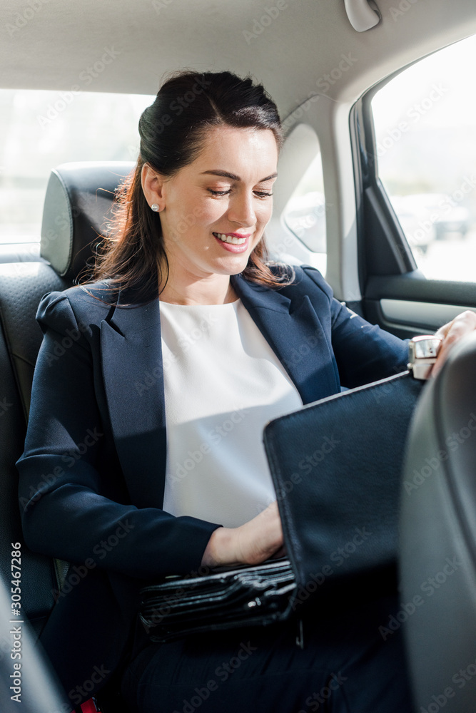 attractive ambassador smiling while sitting in car and touching briefcase