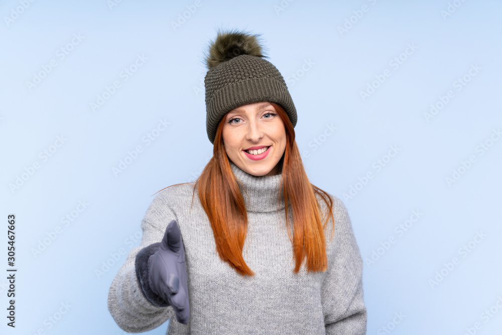 Young redhead Russian woman with winter hat over isolated blue background handshaking after good deal Young redhead Russian woman with winter hat over isolated blue background handshaking after good deal