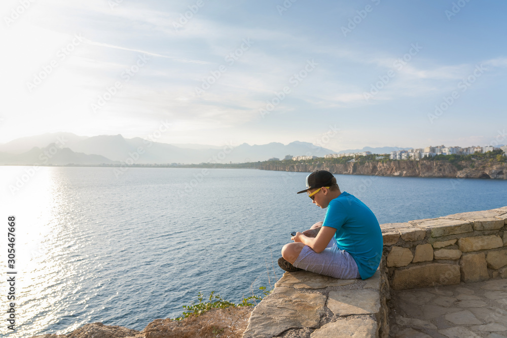Closeup profile view of handsome white kid using modern mobile smartphone sitting in beautiful scenic summer sunny landscape of Turkey, Antalya city. Horizontal color photography. Closeup profile view of handsome white kid using modern mobile smartphone sitting in beautiful scenic summer sunny landscape of Turkey, Antalya city. Horizontal color photography.