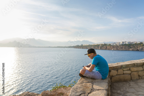 Closeup profile view of handsome white kid using modern mobile smartphone sitting in beautiful scenic summer sunny landscape of Turkey, Antalya city. Horizontal color photography. © Andrii Oleksiienko