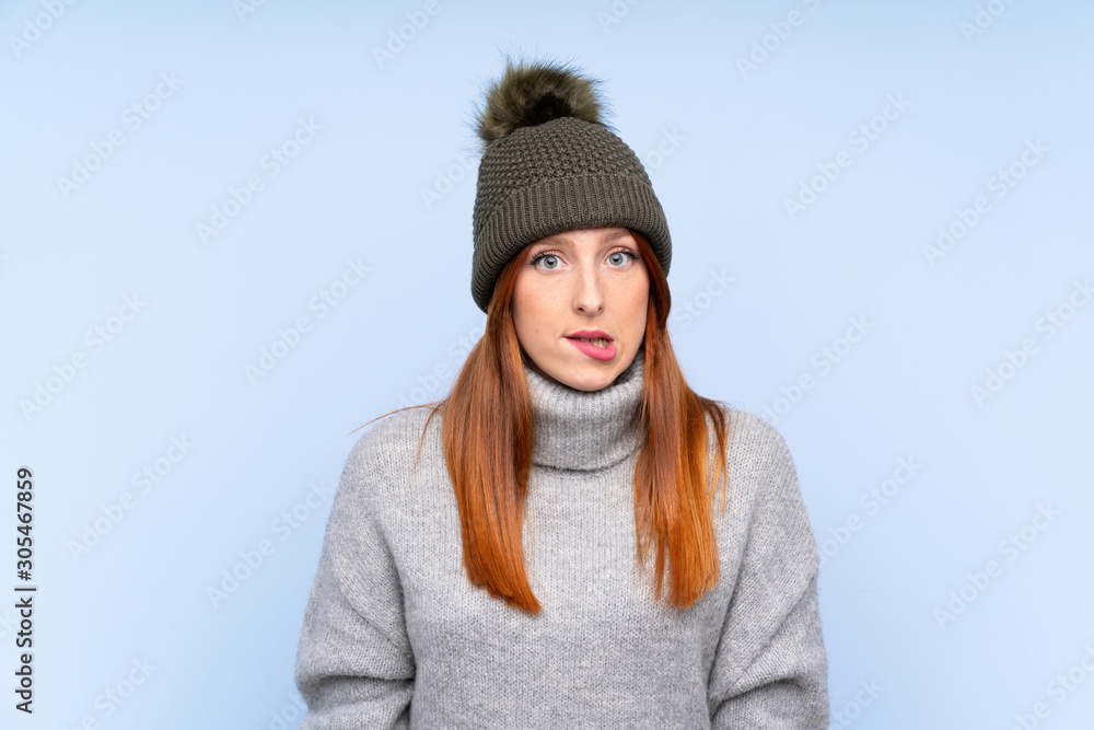 Young redhead Russian woman with winter hat over isolated blue background having doubts and with confuse face expression Young redhead Russian woman with winter hat over isolated blue background having doubts and with confuse face expression
