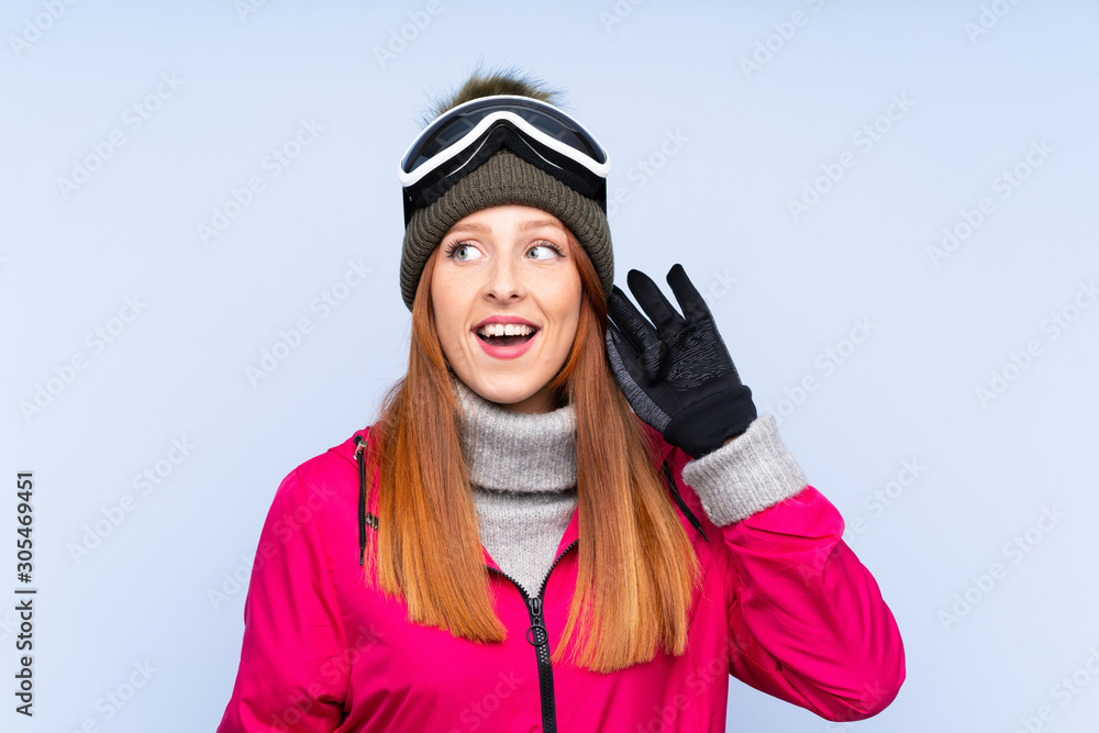 Skier redhead woman with snowboarding glasses over isolated blue wall listening something Skier redhead woman with snowboarding glasses over isolated blue wall listening something