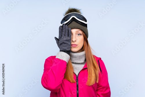 Skier redhead woman with snowboarding glasses over isolated blue wall covering a eye by hand © luismolinero