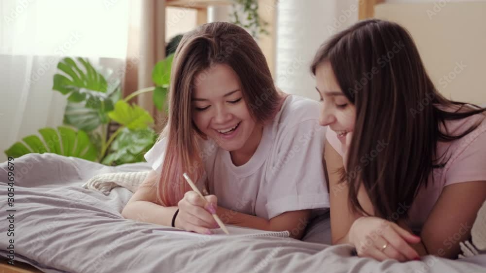 Students lifestyle. Fun homework. Female friends writing essay together lying down on bed. Students lifestyle. Fun homework. Female friends writing essay together lying down on bed.