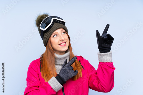 Skier redhead woman with snowboarding glasses over isolated blue wall pointing with the index finger a great idea
