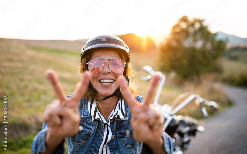 A cheerful senior woman traveller with motorbike in countryside. A cheerful senior woman traveller with motorbike in countryside.