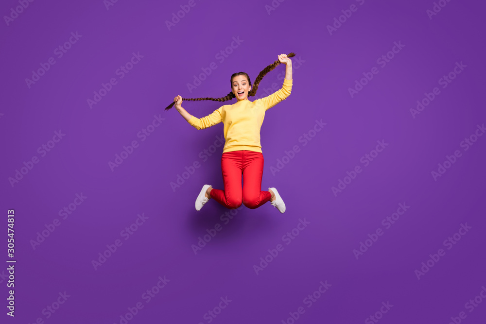 Full body photo of crazy youngster lady jumping high spending best free time holding long braids hands wear casual yellow pullover red trousers isolated purple color background