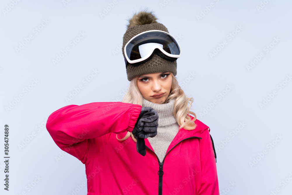 Skier teenager girl with snowboarding glasses over isolated blue background showing thumb down sign Skier teenager girl with snowboarding glasses over isolated blue background showing thumb down sign