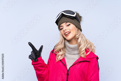Skier teenager girl with snowboarding glasses over isolated blue background pointing finger to the side