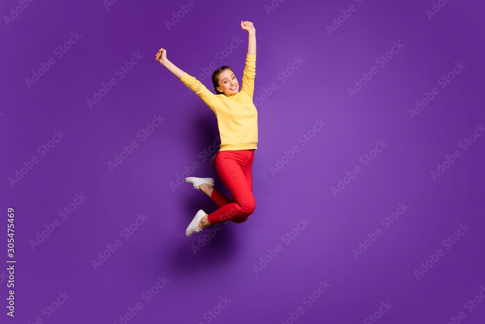 Full length photo of pretty teen lady jumping high celebrating vacation time raising hands up wear casual yellow pullover red pants isolated purple color background