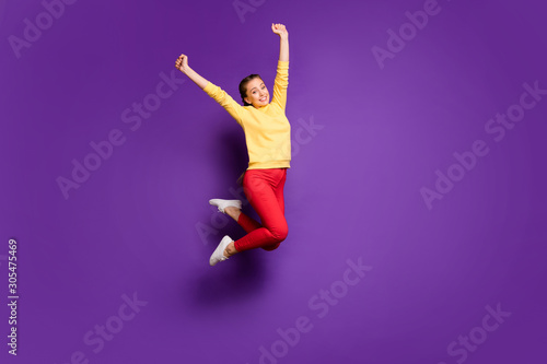 Full length photo of pretty teen lady jumping high celebrating vacation time raising hands up wear casual yellow pullover red pants isolated purple color background