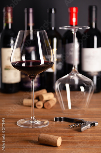 Crystal shiny glass of red wine, bottles, corkscrew, opener, sommelier knife, transparent decanter, corks on wooden table. Closeup, vertical, selective focus side view. Concept wine magazine photo