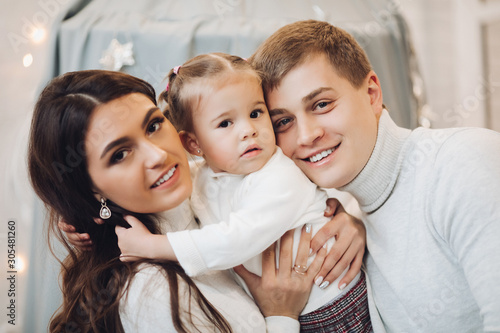 Stock portrait of attractive Caucasian family hugging and smiling at camera with daughter on hands. Brunette mother and father with adorable daughter smiling at camera.