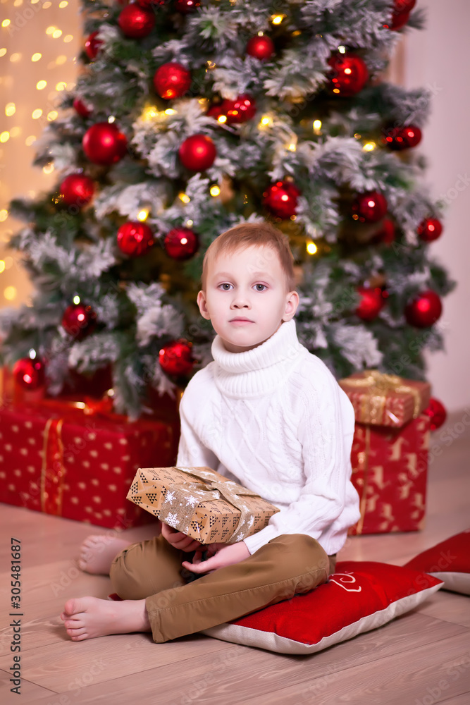Child in white knitted sweater holding a Christmas gift box in a hands and sitting under Christmas tree