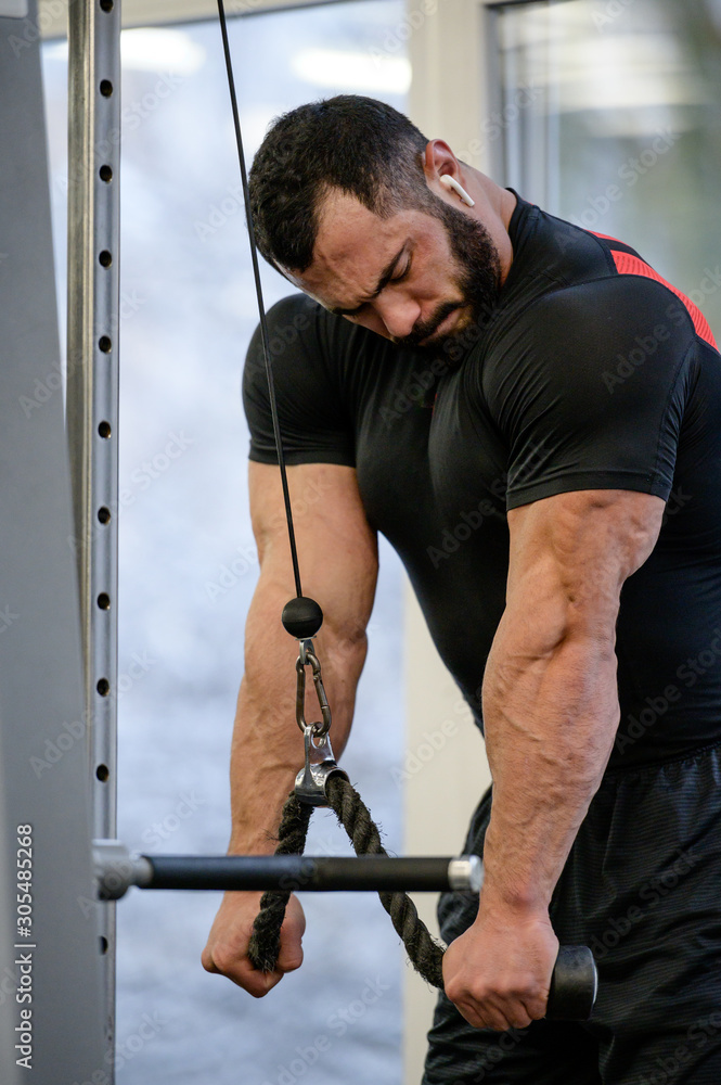 strong healthy young bearded sport man in black sportswear and wireless headset during workout for muscle triceps pulling heavy weight using rope strong healthy young bearded sport man in black sportswear and wireless headset during workout for muscle triceps pulling heavy weight using rope