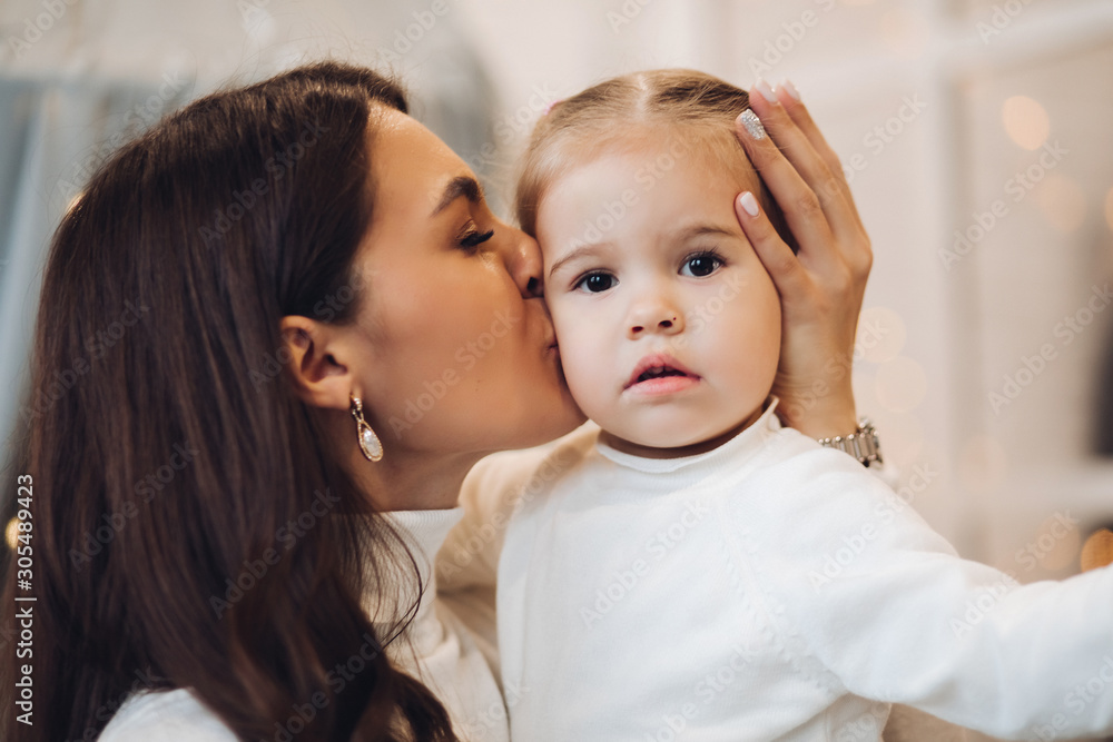Stock photo of loving brunette mother holding her little cute daughter on arms and kissing her cheek. They are in room decorated for Christmas.