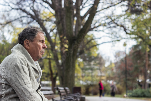 Portrait of a pensive senior man sitting on the bench, in the public park, outdoors. Old man relaxing outdoors and looking away. Portrait of elderly man enjoying retirement