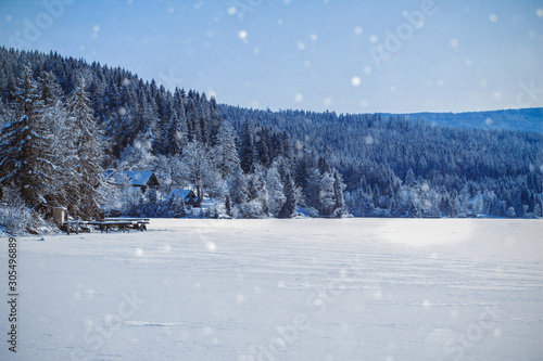 Winter am See-Schneeflocken fallen auf einen zugefrorenen See mit Wald und kleinen Holzhütten drum herum-Weihnachtspostkarte