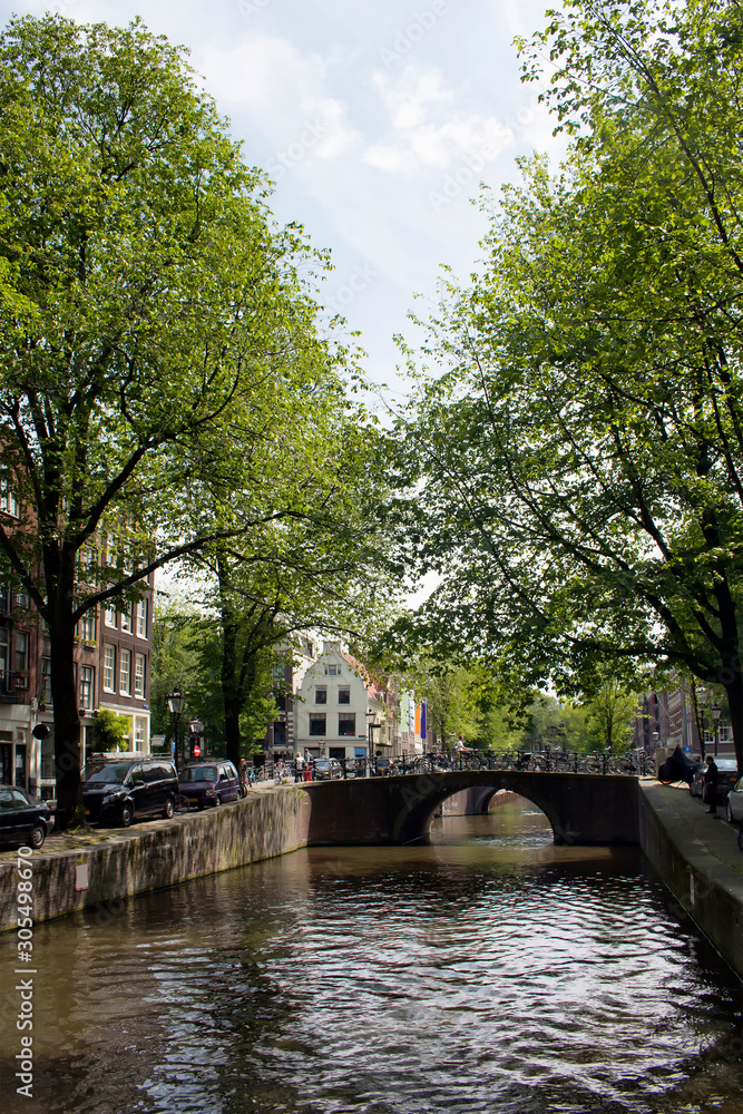 View of canal, stone bridge, trees, parked cars in Amsterdam. It is a sunny summer day.