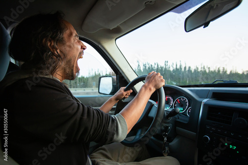 Side view photo of adult middle aged male with long dark hair driving his truck car through nature scenes while yawning and being very tired  © Valmedia