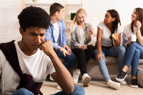Upset bullied guy sitting alone, excluded by bad friends photo