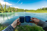 Selective focus of the hands of two people clinching their cups of coffee and tea with in the background a crystal clear lake and trees