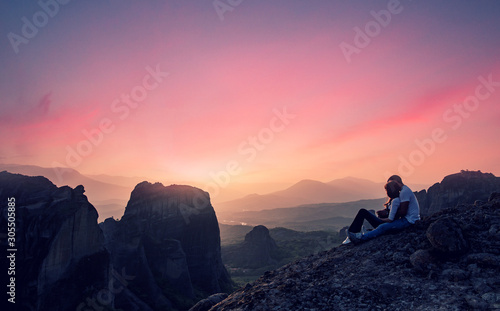 Lovely Couple sitting on rock in the evening, beautiful dusk. Wonderful Panoramic view of the rocks and monasteries of Meteora, Greece. Mysterious colorful sky, during sunset. Awesome Nature Landscape