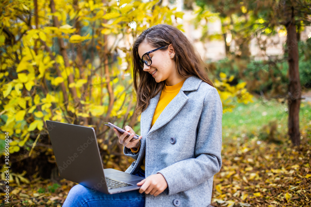Young pretty woman working on laptop while use mobile phone sitting on the bench in autumn park Young pretty woman working on laptop while use mobile phone sitting on the bench in autumn park