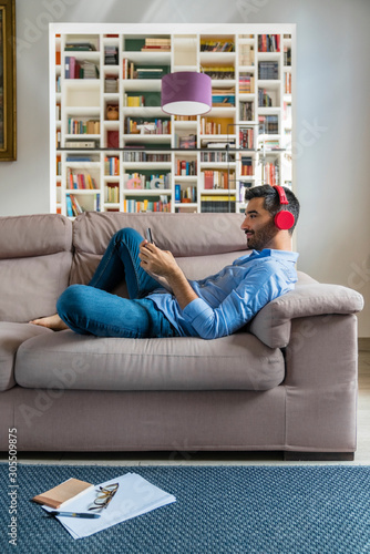 Smiling young man lying on the couch at home using smartphone and wireless headphones