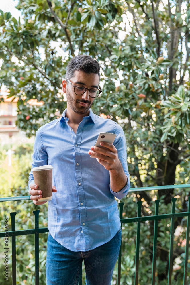 Portrait of young man standing on balcony with coffee to go looking at cell phone Portrait of young man standing on balcony with coffee to go looking at cell phone