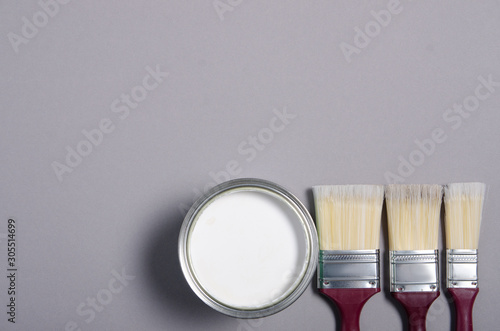 Open can with white paint and brush on gray background, top view. Space for text