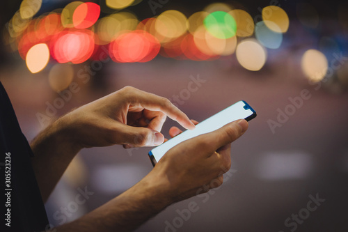 Cropped view of male fingers typing text message on blank display of modern smartphone on bokeh background. Man's hands holding mobile phone and chatting online in social networks.