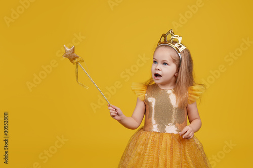 Fotografia Joyful little girl with long hair in a tulle golden dress and princess crown holding a magic wand  on yellow background