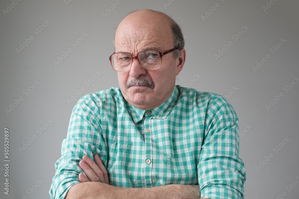 Angry senior man in glasses crossing arms looking annoyed at camera. He does not trust the news he is told. Studio shot Angry senior man in glasses crossing arms looking annoyed at camera. He does not trust the news he is told. Studio shot