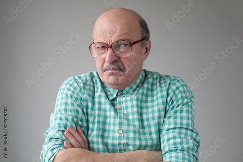 Angry senior man in glasses crossing arms looking annoyed at camera. He does not trust the news he is told. Studio shot