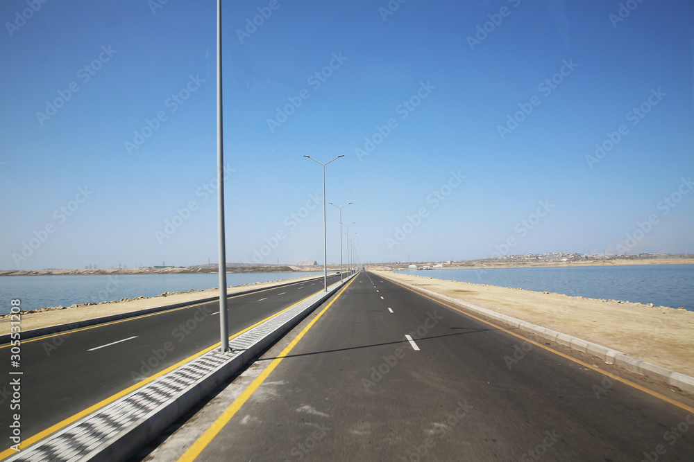 road under a blue sky with focus to the center white line . asphalt car road .wallpaper landscape .sea and lake road .light poles . Empty road white cloudy weather.