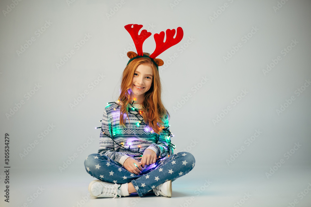 Beautiful little girl in a garland sitting widely smiles at the camera in the New Year The red-haired lady wrapped a garland to create festive mood A teenager is sitting on the floor before Christmas Beautiful little girl in a garland sitting widely smiles at the camera in the New Year The red-haired lady wrapped a garland to create festive mood A teenager is sitting on the floor before Christmas