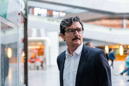 Bewildered handsome aged male entrepreneur in business style clothing and eyeglasses looking away on blurred background at shopping center photo