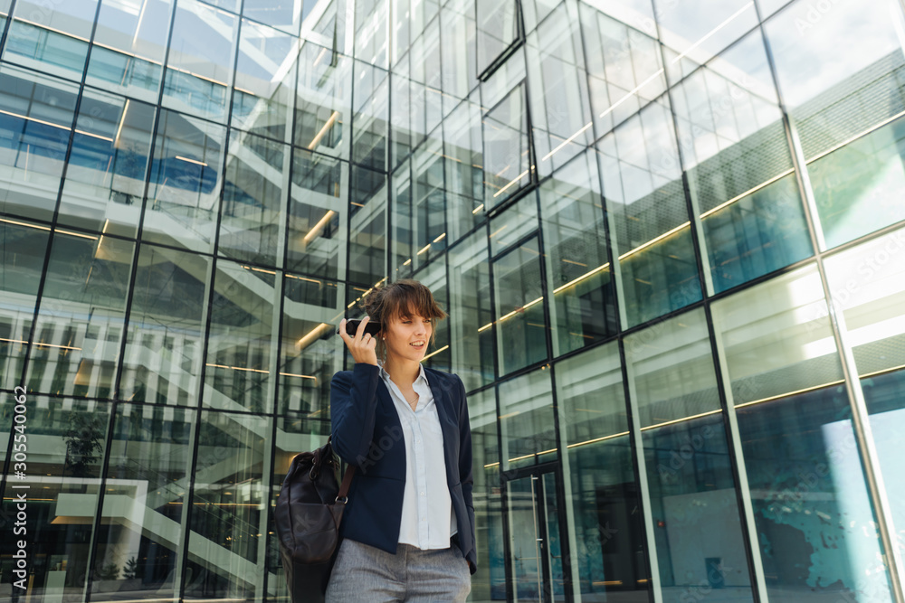 Low angle of happy businesswoman holding hand in pocket and using smartphone while standing outside business center on city street Low angle of happy businesswoman holding hand in pocket and using smartphone while standing outside business center on city street